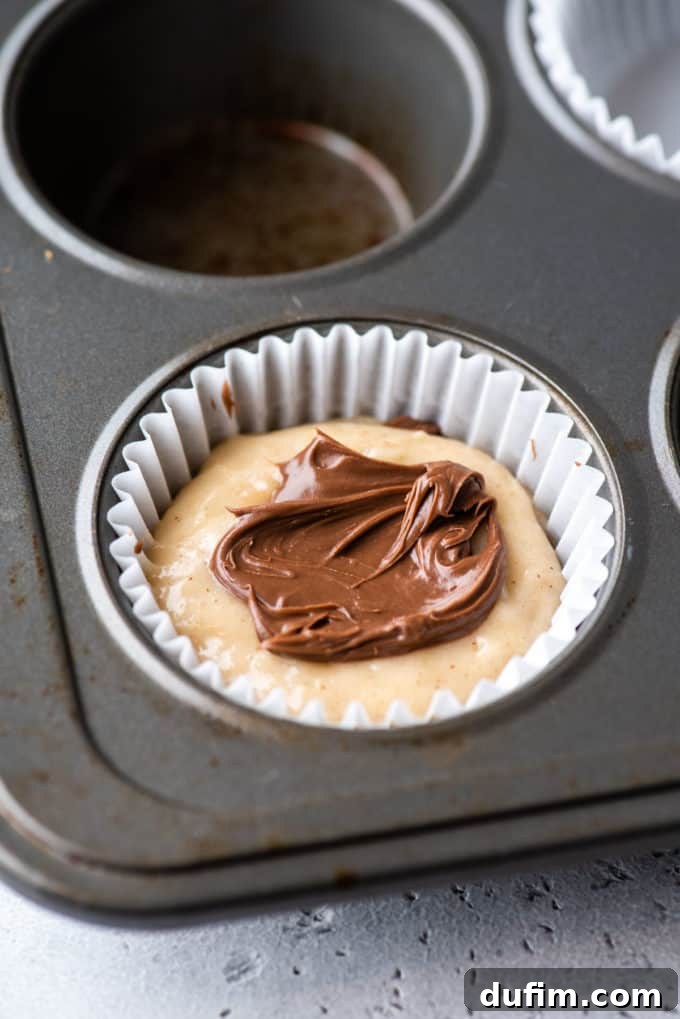 A close-up of a muffin pan with a white muffin liner in the corner, showing carefully layered muffin batter alternating with creamy Nutella inside the liner.