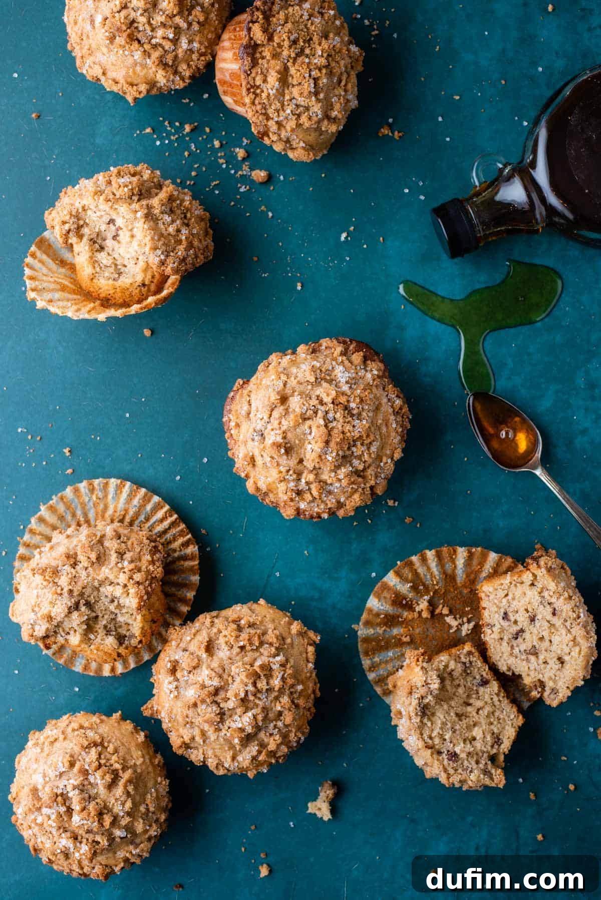 Golden Maple Pecan Muffins 2 Several golden-brown maple pecan muffins arranged artfully on a dark teal surface. Some are upright, showcasing their domed tops, while others are on their side, revealing their moist interiors. A bottle of rich maple syrup and a spoon glistening with syrup are placed nearby, enhancing the theme.