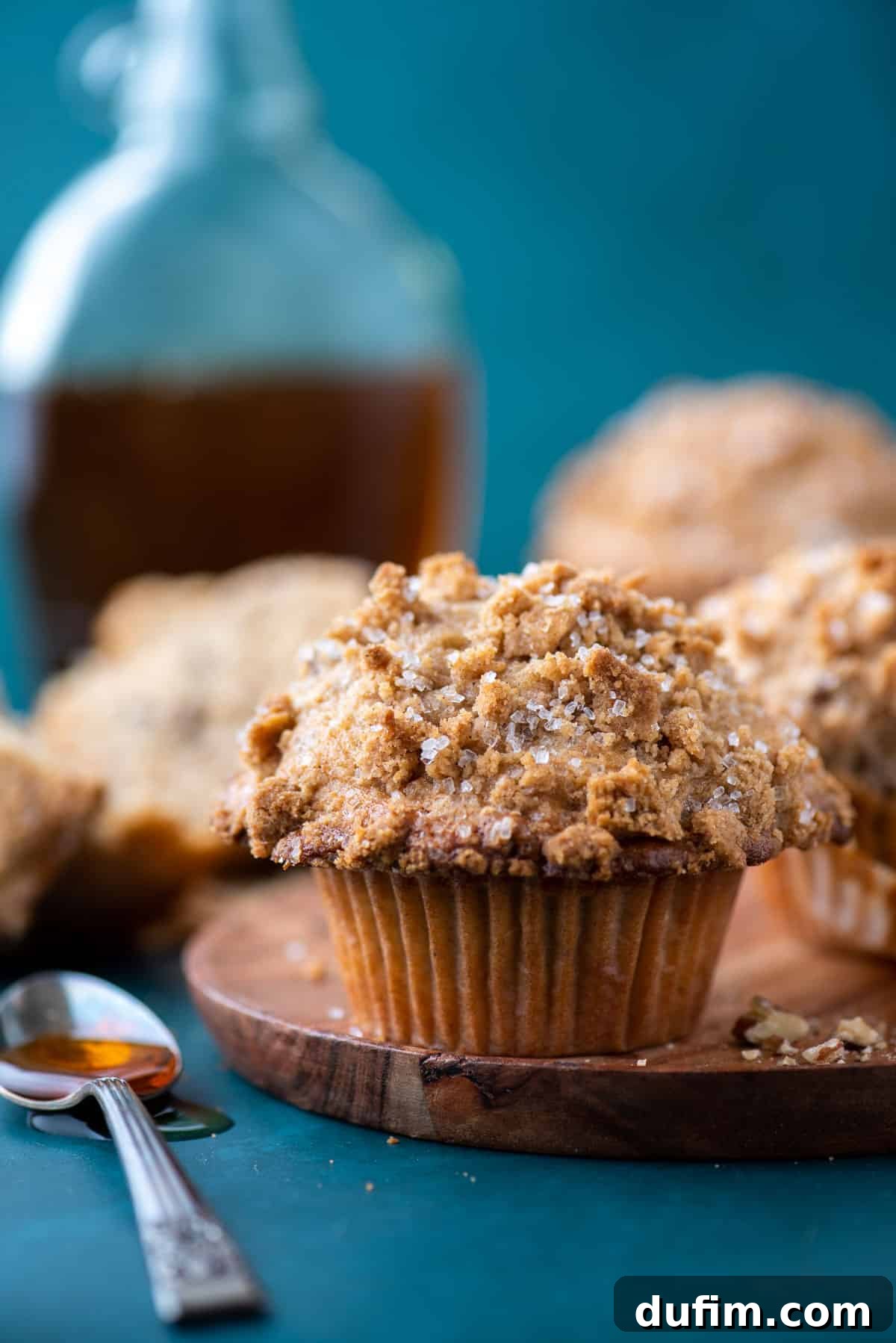 Golden Maple Pecan Muffins 3 A single, beautifully baked maple pecan muffin rests invitingly on a rustic wood plate, placed atop a contrasting dark teal surface. More muffins are softly blurred in the background, creating a tempting scene. A spoon with glistening maple syrup beside it adds a touch of sweetness and visual appeal.
