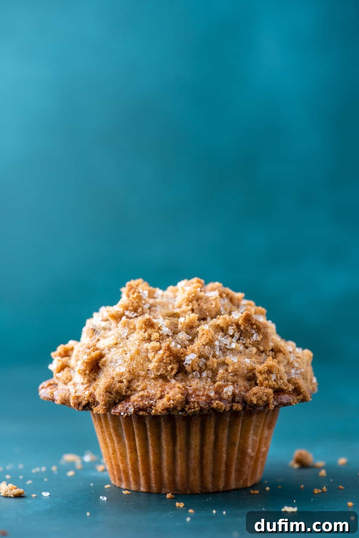 Golden Maple Pecan Muffins 4 A perfectly baked maple pecan muffin rests delicately on a dark teal surface, surrounded by delightful, golden-brown crumbs from its delicious topping. The muffin's impressive domed top and visible pecan pieces promise a truly delicious and satisfying bite.