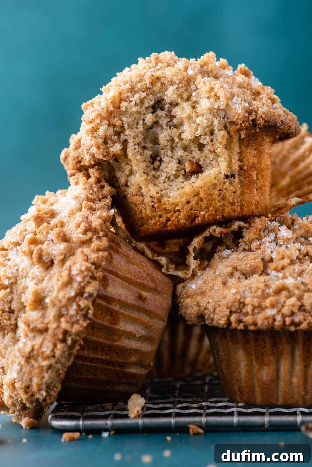 Golden Maple Pecan Muffins 5 A generous pile of freshly baked maple pecan muffins, some with delicious crumb topping, cooling invitingly on a wire rack. The rack is placed on a dark teal surface, with the top muffin visibly missing a bite, hinting at its soft, delicious interior.
