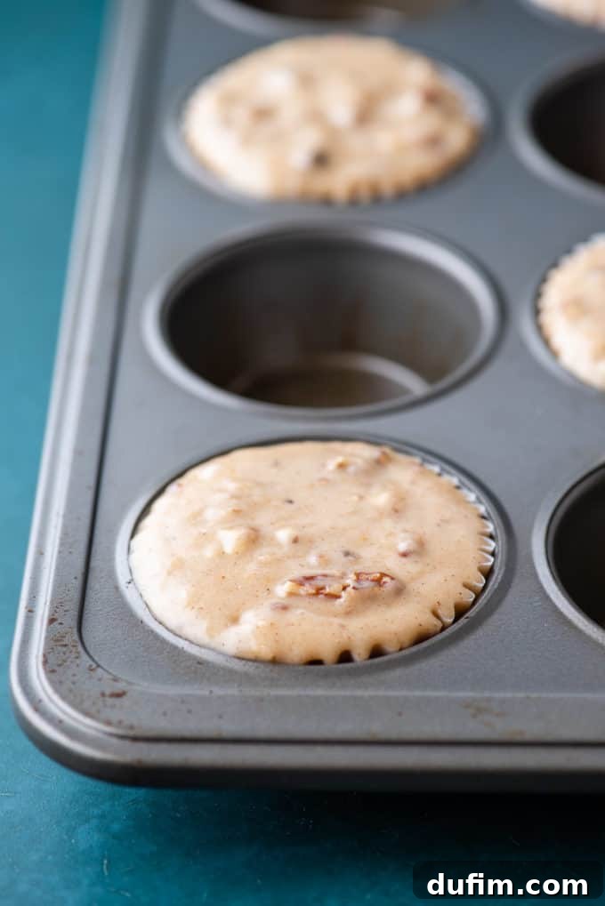 Golden Maple Pecan Muffins 7 A muffin pan resting on a dark teal surface, meticulously prepared with maple pecan muffin batter strategically placed in every other muffin well. This deliberate spacing is crucial for achieving large, impressive, and perfectly domed muffin tops.