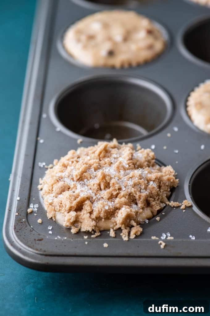 Golden Maple Pecan Muffins 8 A muffin pan on a dark teal surface, now featuring maple pecan muffins in every other hole, generously adorned with crumb topping and a delicate sprinkle of sparkling sanding sugar, all ready for the oven.