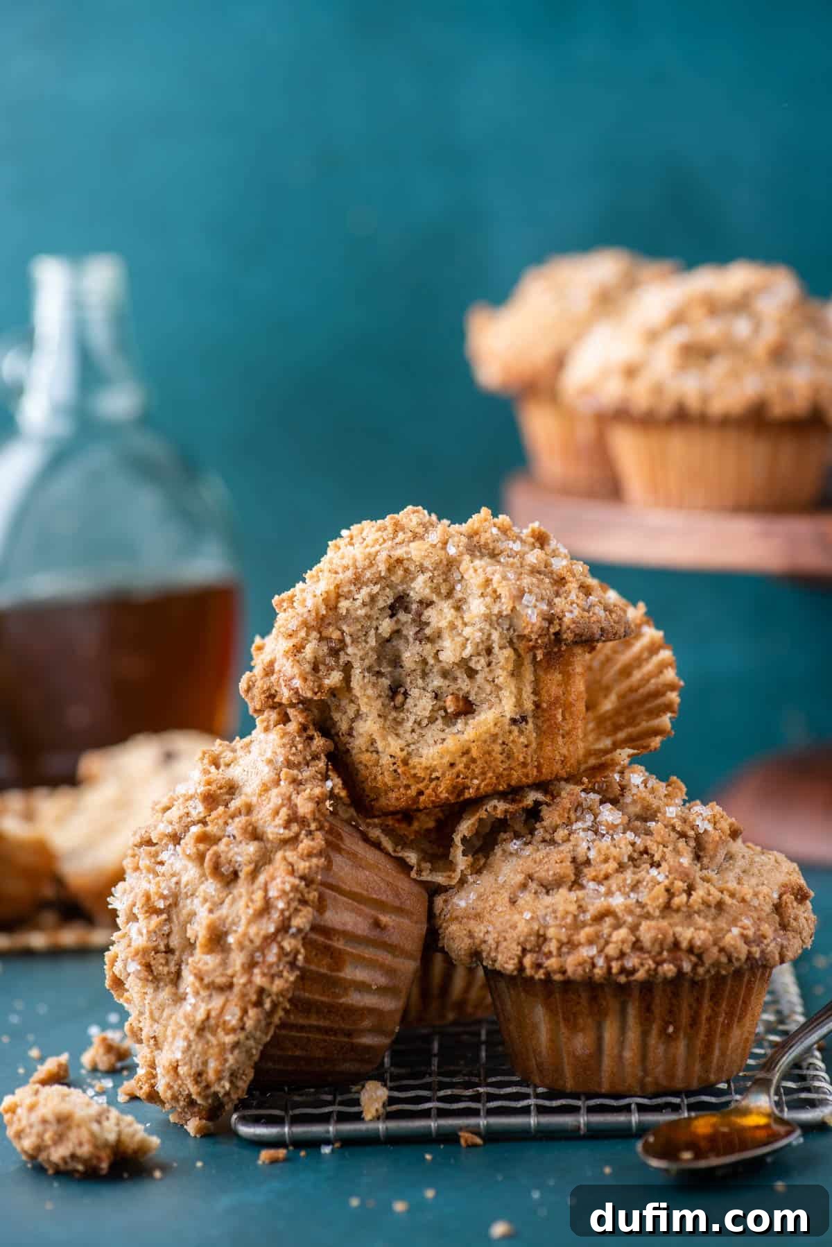 Golden Maple Pecan Muffins 10 A delightful pile of golden-brown maple pecan muffins resting atop a wire rack, with the top muffin showing a bite taken out, revealing its moist and tender interior. A spoon full of glistening pure maple syrup rests nearby, and more muffins and maple syrup bottles are softly blurred in the background, creating an inviting culinary scene.