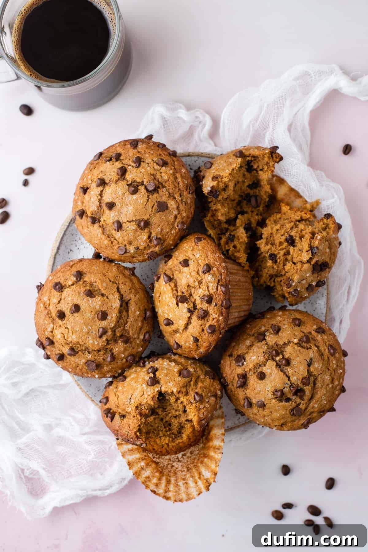 Café Mocha Chip Muffins 2 A plate full of golden-brown cappuccino chip muffins on a white cheesecloth, surrounded by coffee beans and a clear glass cup of coffee.