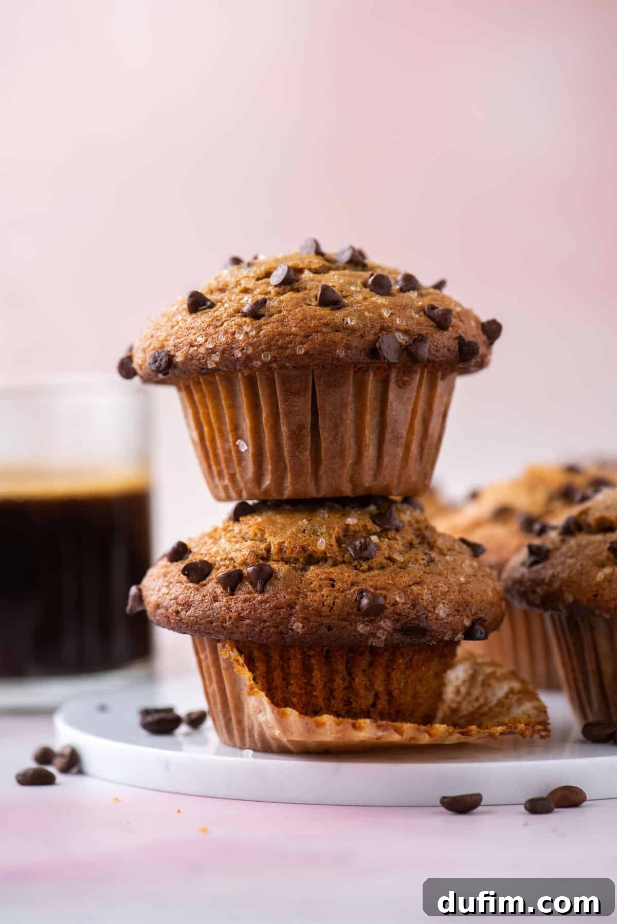 Café Mocha Chip Muffins 4 A stack of two cappuccino chip muffins, garnished with coffee beans. More muffins and a cup of coffee are visible in the soft background.