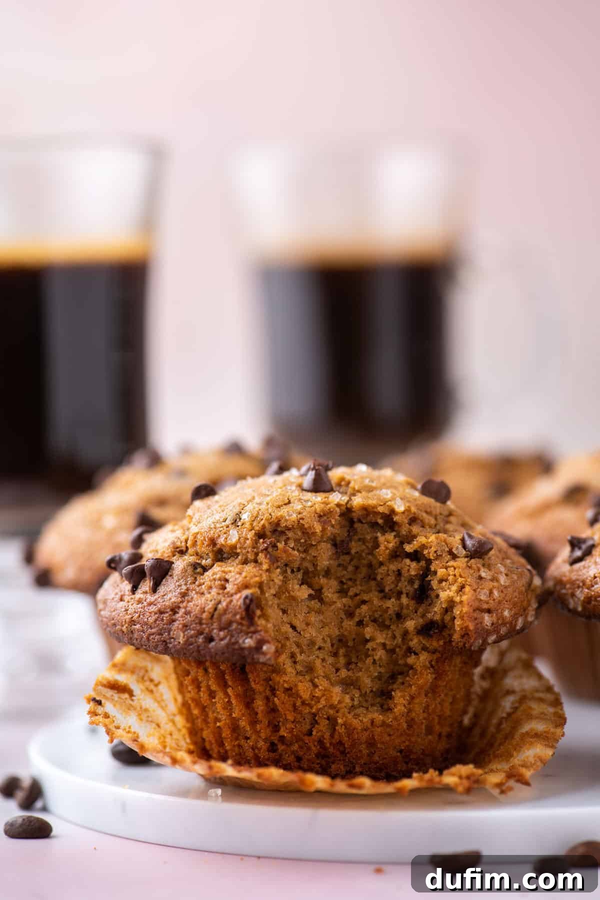 Café Mocha Chip Muffins 5 A cappuccino chip muffin on a white plate, with a bite taken out, sitting in its open muffin liner. Coffee beans are scattered around, with more muffins and two cups of coffee in the background.