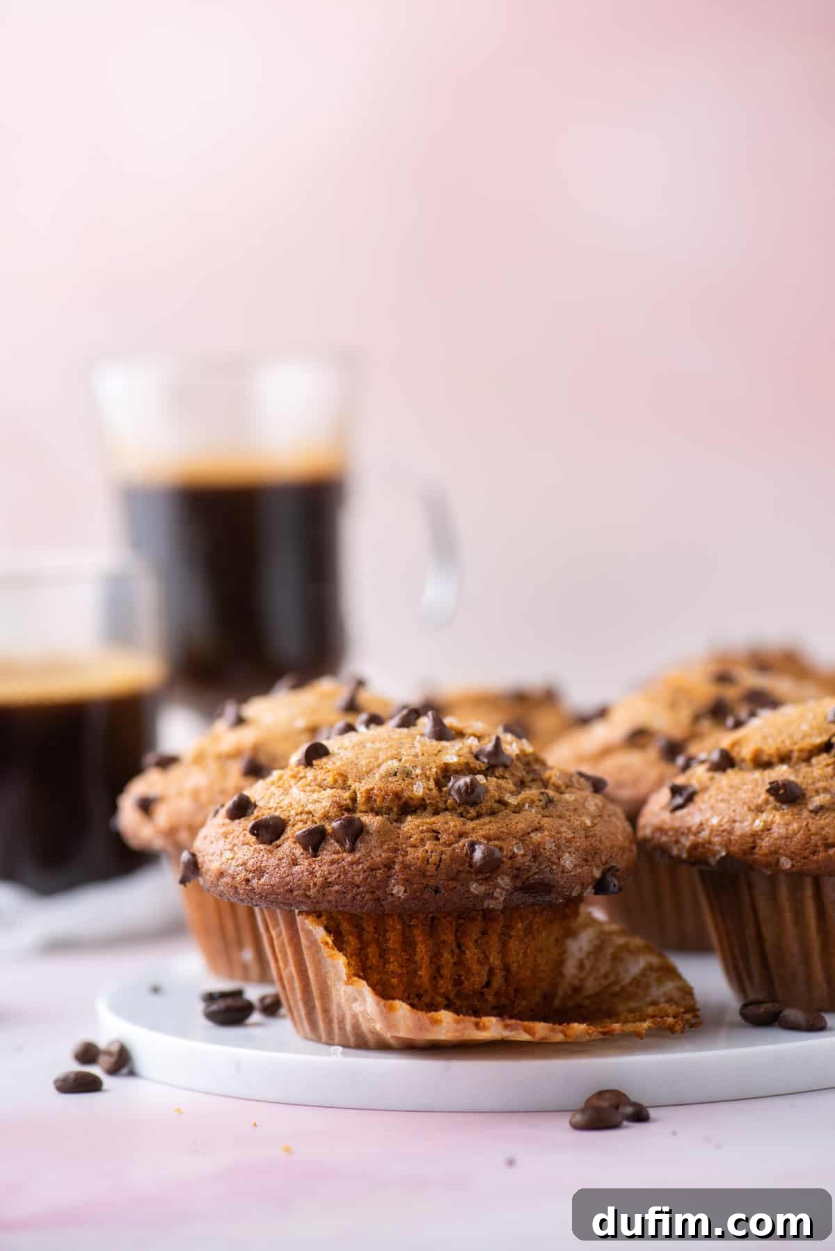 Café Mocha Chip Muffins 8 Cappuccino chip muffins on a white plate, nestled in their open muffin liners. Coffee beans are scattered around, with more muffins and two cups of coffee in the background.