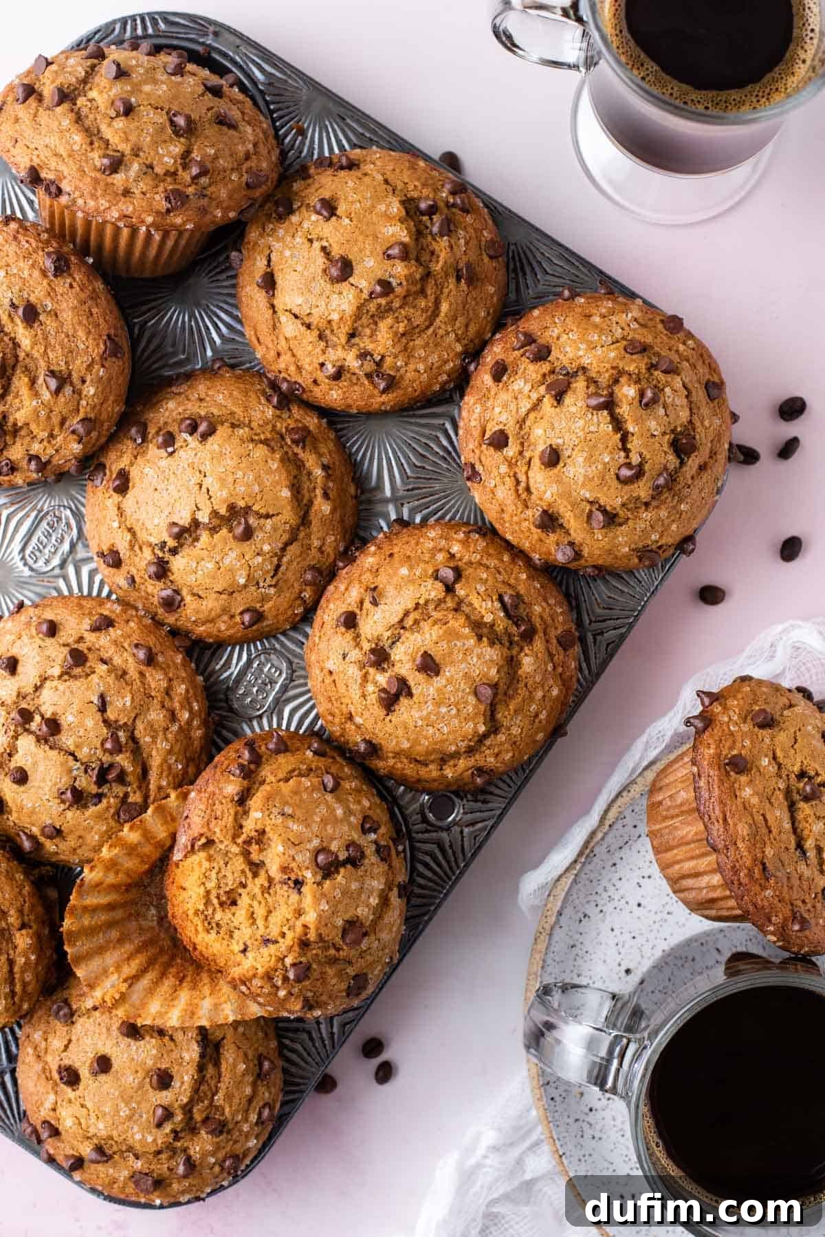 Café Mocha Chip Muffins 9 An overhead view of a muffin pan full of cappuccino chip muffins, surrounded by coffee beans. A cup of coffee and a plate with a muffin are beside it.