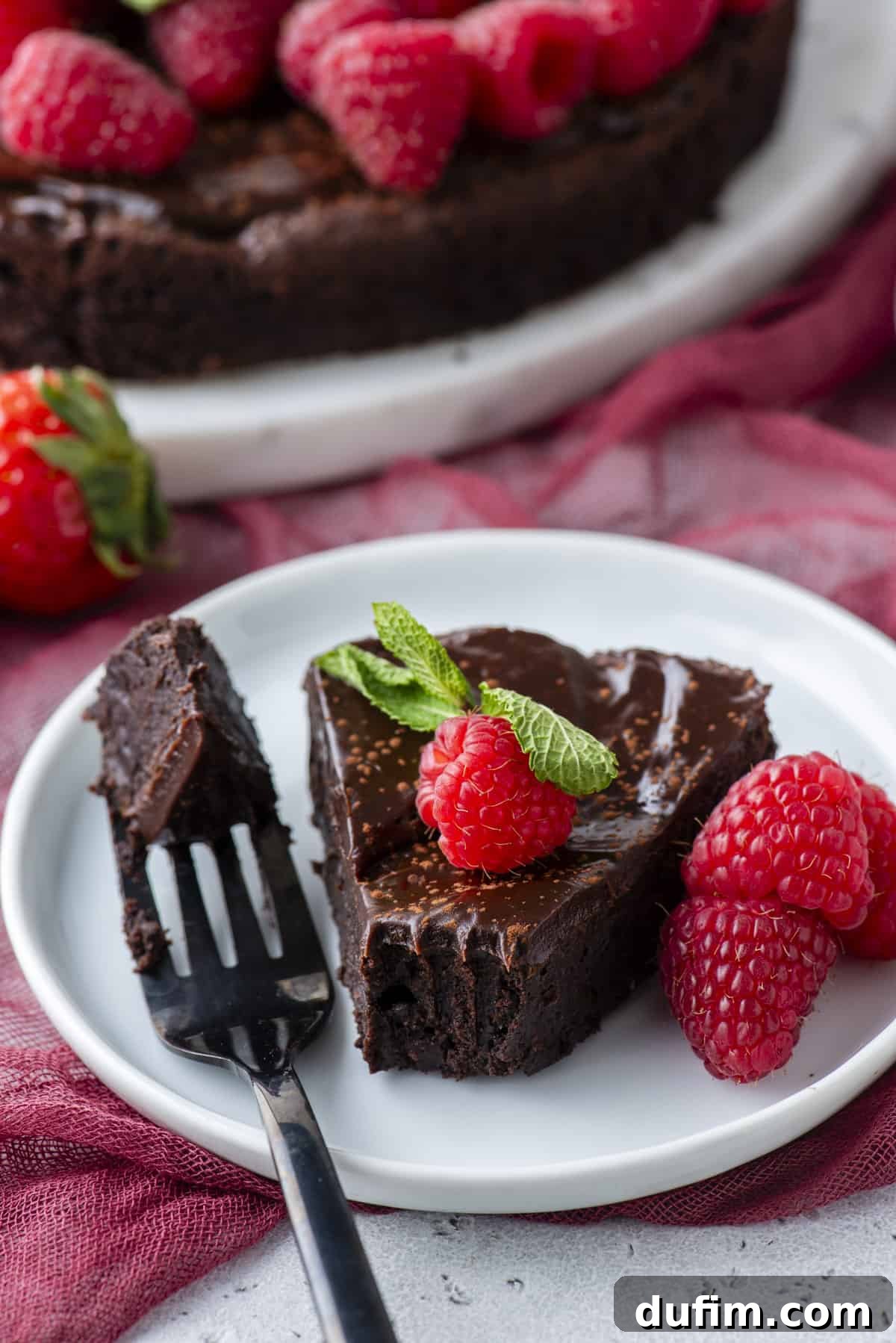 Decadent Flourless Chocolate Cake 17 a slice of flourless chocolate cake on a white plate topped with a fresh raspberry and mint, with a fork laying beside it with one bite on it, more fresh raspberries beside it and more cake in the background