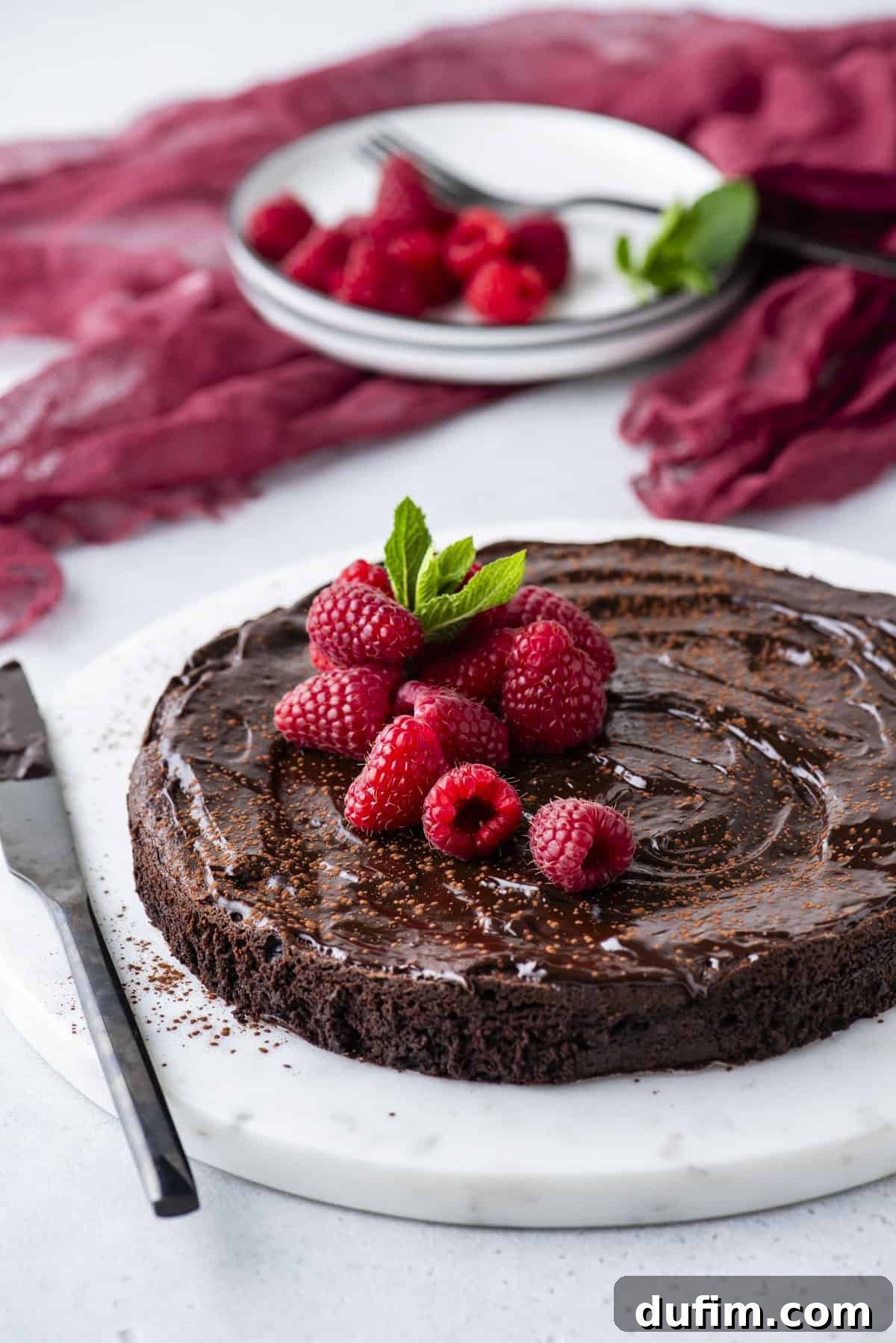 Decadent Flourless Chocolate Cake 18 a flourless chocolate cake on a round white serving platter topped with fresh raspberries and mint leaves, with a knife covered in chocolate laying beside it, a red cloth and a bowl of fresh raspberries and mint in the background