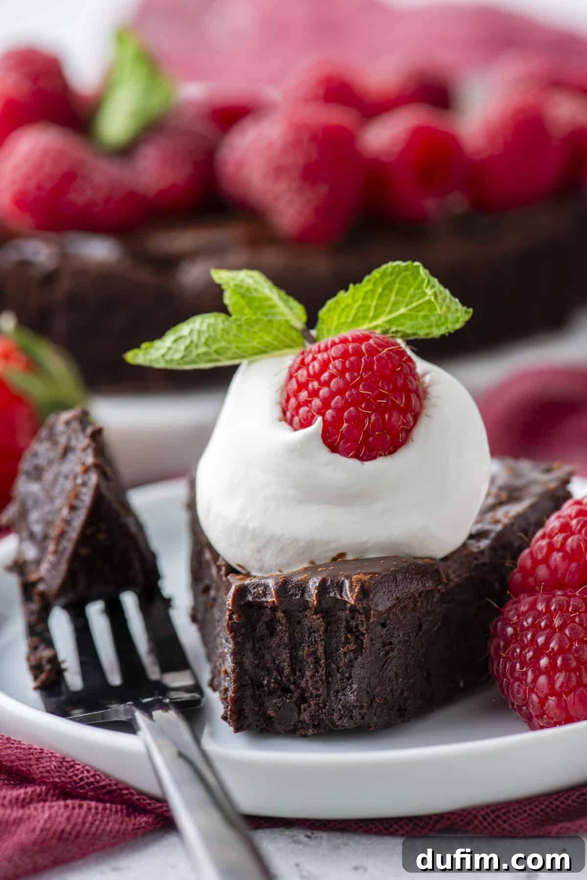 Decadent Flourless Chocolate Cake 4 a slice of flourless chocolate cake on a white plate, topped with whipped cream, a fresh raspberry and mint, with a fork laying beside it with one bite on it, more fresh raspberries beside it and more cake in the background