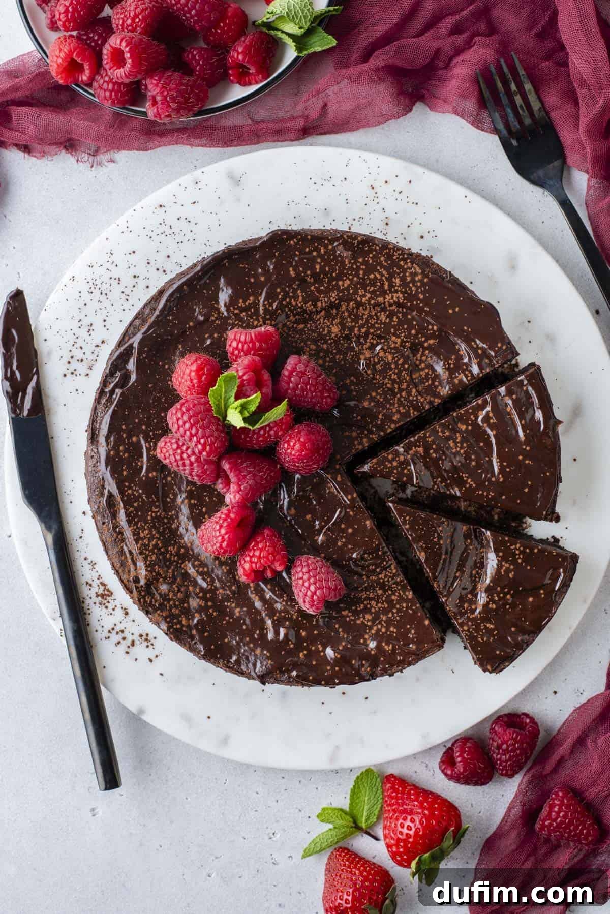 Decadent Flourless Chocolate Cake 5 overhead shot of a full flourless chocolate cake with two slices cut, sitting on a round white serving platter, topped with fresh raspberries and mint leaves, with a knife covered in chocolate laying beside it, red clothes and more raspberries and fresh strawberries surrounding it