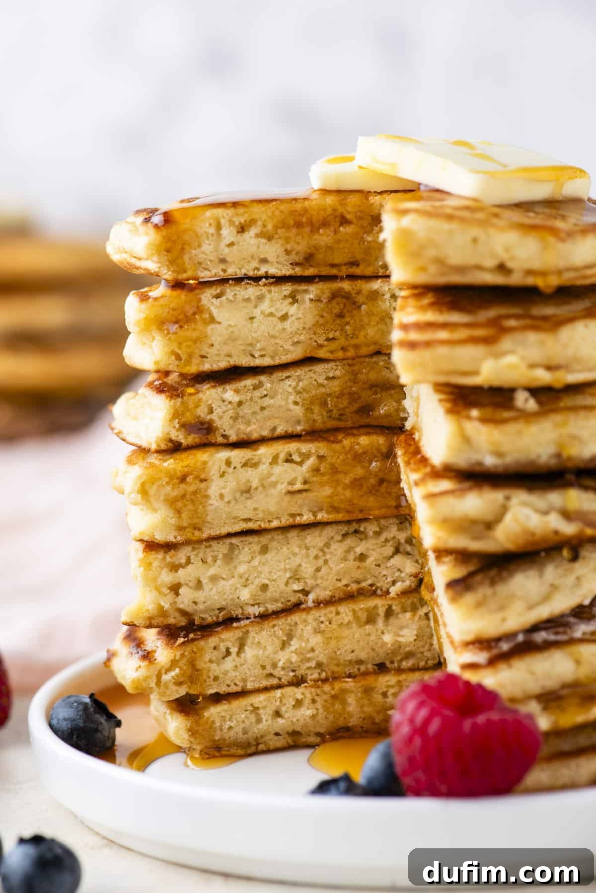 A tall stack of fluffy golden pancakes with a triangular portion cut out, generously drizzled with rich maple syrup, adorned with a pat of butter, and surrounded by vibrant fresh berries on a pristine white plate.