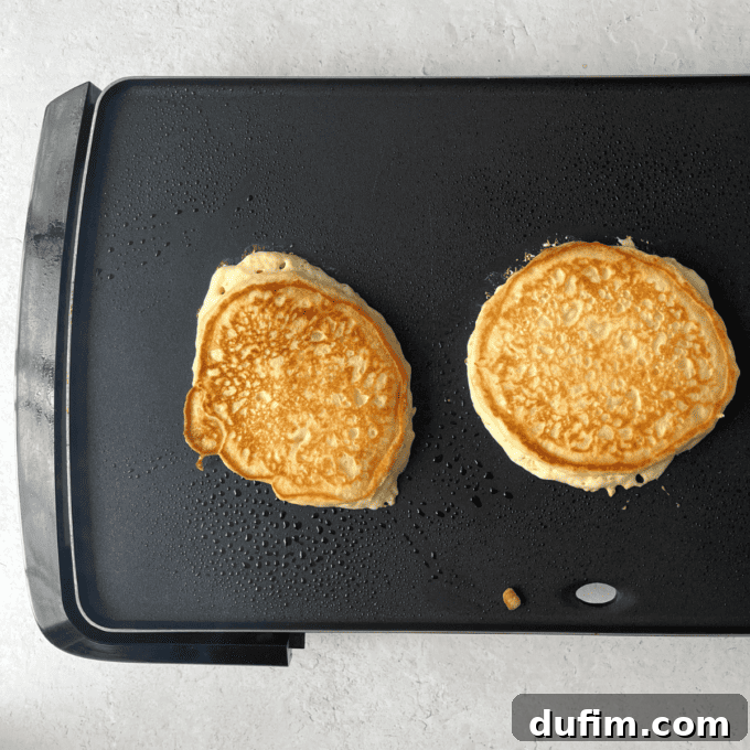 Two perfectly golden-brown pancakes are cooking side-by-side on a flat-top griddle, with steam gently rising, showcasing the ideal cooking stage before serving.