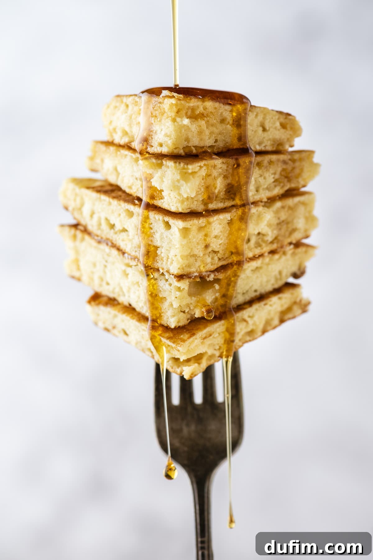 A delectable stack of pancake slices, artfully impaled and held vertically on a fork, with glistening syrup cascading down the sides, capturing the irresistible texture and appeal of freshly made pancakes.