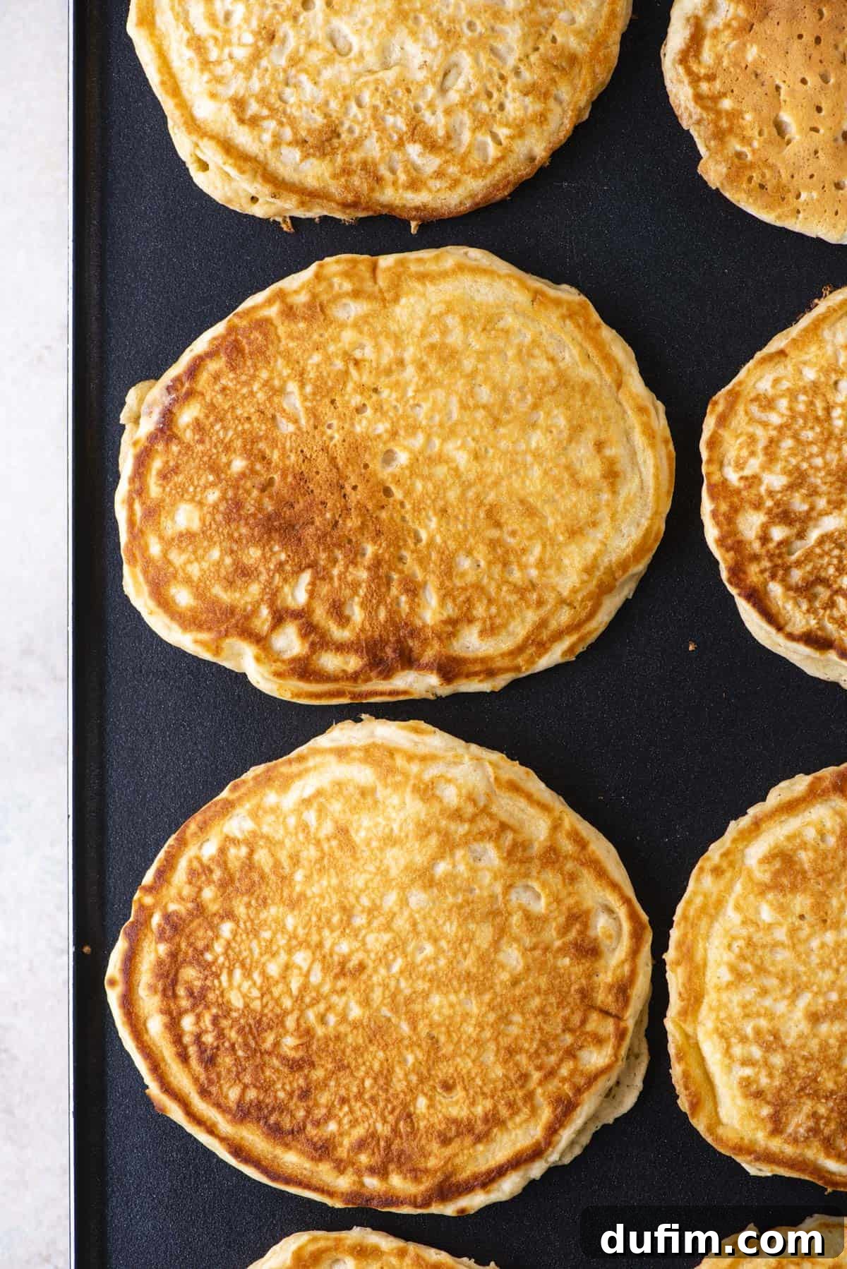 Multiple rows of perfectly golden-brown pancakes are cooking simultaneously on a large griddle, showcasing efficient preparation for a big breakfast or brunch.