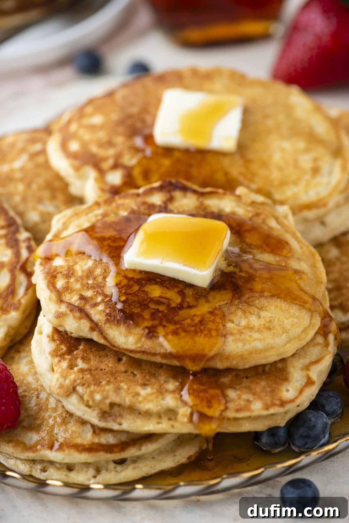 A sumptuous plate piled high with golden-brown pancakes, topped with a melting pat of butter and glistening with syrup. Fresh, colorful berries are scattered generously around the base, creating a vibrant and appealing breakfast scene with more pancakes and berries blurred in the background.