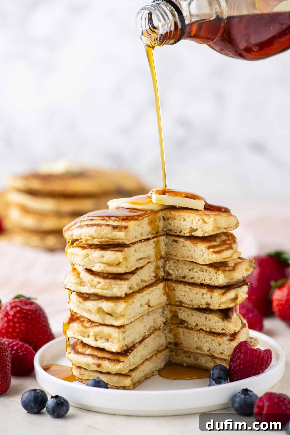 A visually striking scene: a tall stack of beautifully cooked pancakes with a triangular section expertly removed, being drenched in a continuous stream of golden maple syrup. A luxurious pat of butter sits atop the stack, slowly melting, while an array of glistening fresh berries artfully encircles the pancakes on a crisp white plate, creating an inviting breakfast tableau.