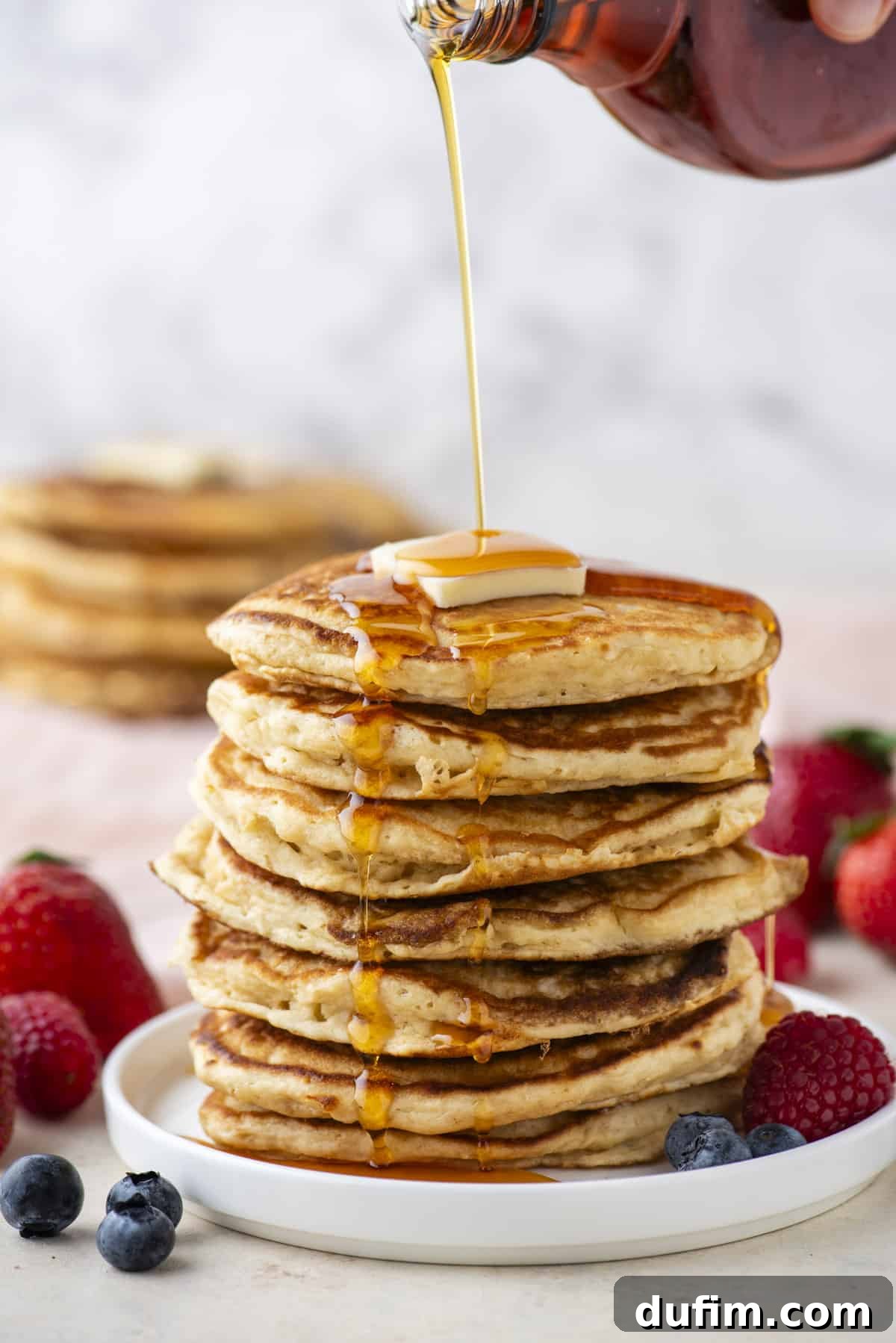 A magnificent tall stack of fluffy homemade pancakes resting on a white plate, elegantly drizzled with golden maple syrup and crowned with a melting pat of butter. A scattering of fresh, vibrant berries provides a colorful accent, while more pancakes are visible in the soft background, creating a mouthwatering breakfast scene.