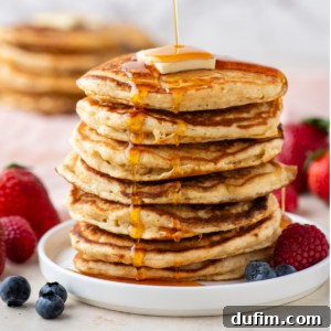 A tall stack of fluffy golden pancakes on a white plate, elegantly drizzled with syrup, topped with a pat of butter, and surrounded by fresh berries. More pancakes are visible in the background.