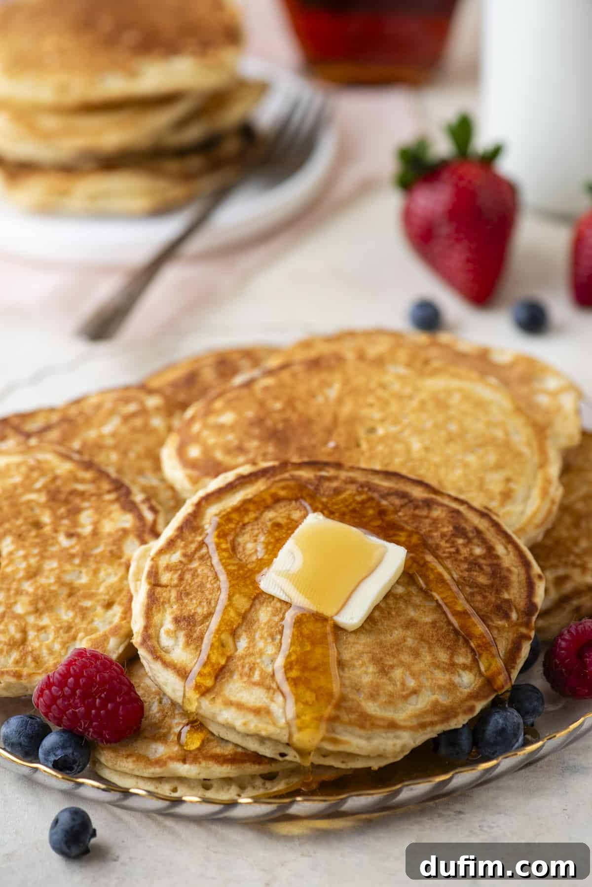 A beautiful arrangement of fluffy pancakes piled high on a white plate, with one pancake elegantly topped with melted butter and glistening syrup. Fresh, vibrant berries artfully surround the stack, with more pancakes and berries softly blurred in the background, hinting at a bountiful breakfast spread.
