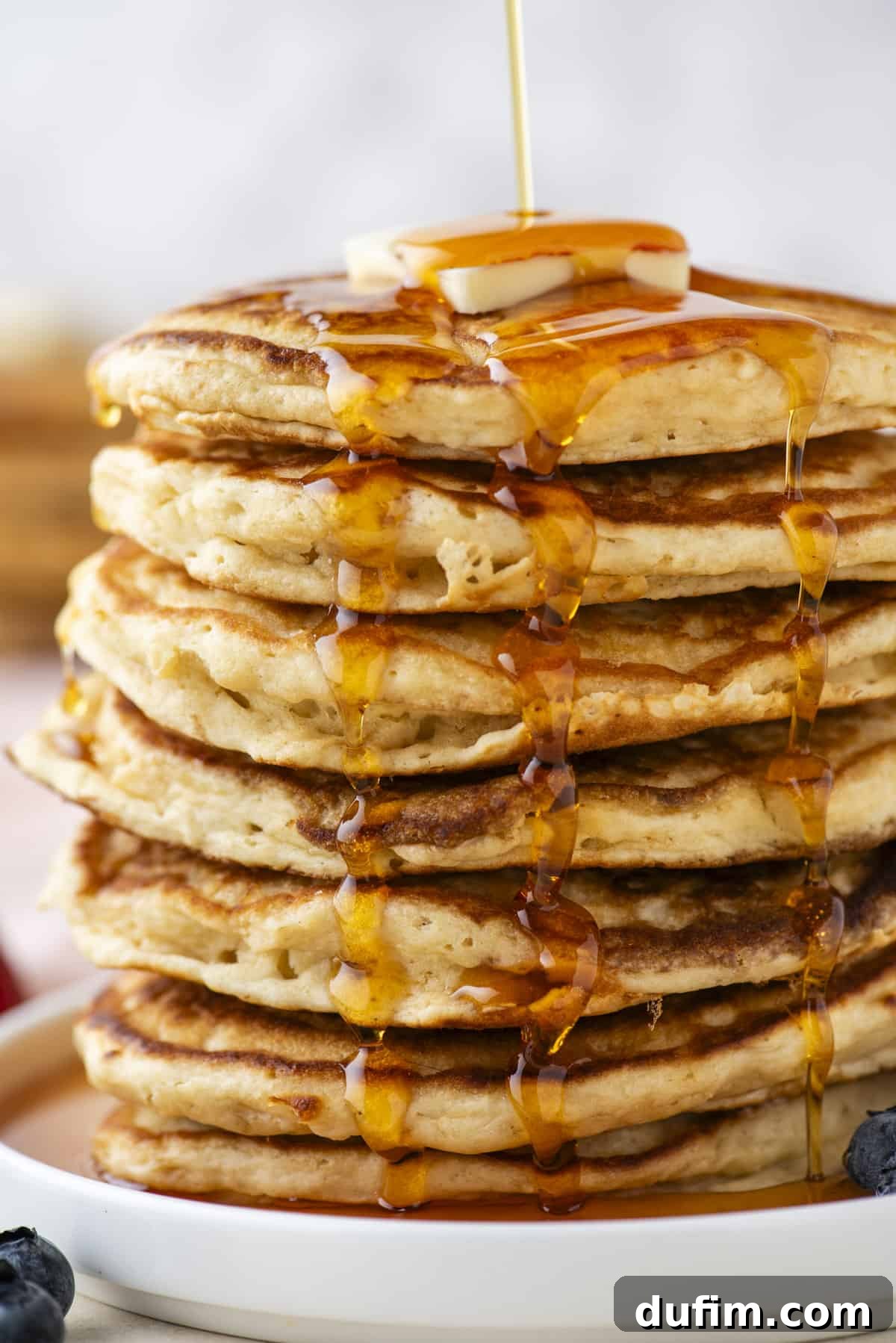 A stunning close-up of a generously tall stack of fluffy pancakes on a pure white plate, being gracefully drizzled with a stream of golden syrup. A perfectly placed pat of butter rests on top, melting into the warmth, while an abundance of fresh, ripe berries adds a vibrant pop of color and freshness around the base.