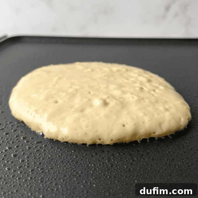 A single pancake cooks on a hot griddle, with numerous small bubbles actively forming on its surface, indicating it's almost ready to be flipped to achieve perfect golden-brown on both sides.