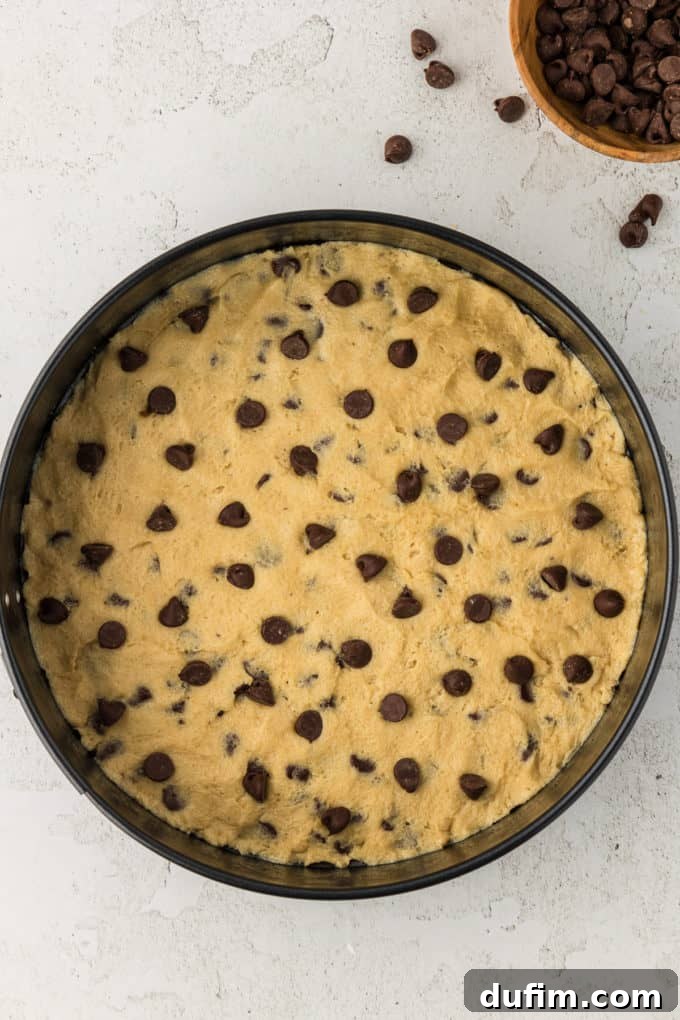 Prepared cookie cake batter evenly spread in a round cake pan, with a small bowl of extra chocolate chips next to it for topping.