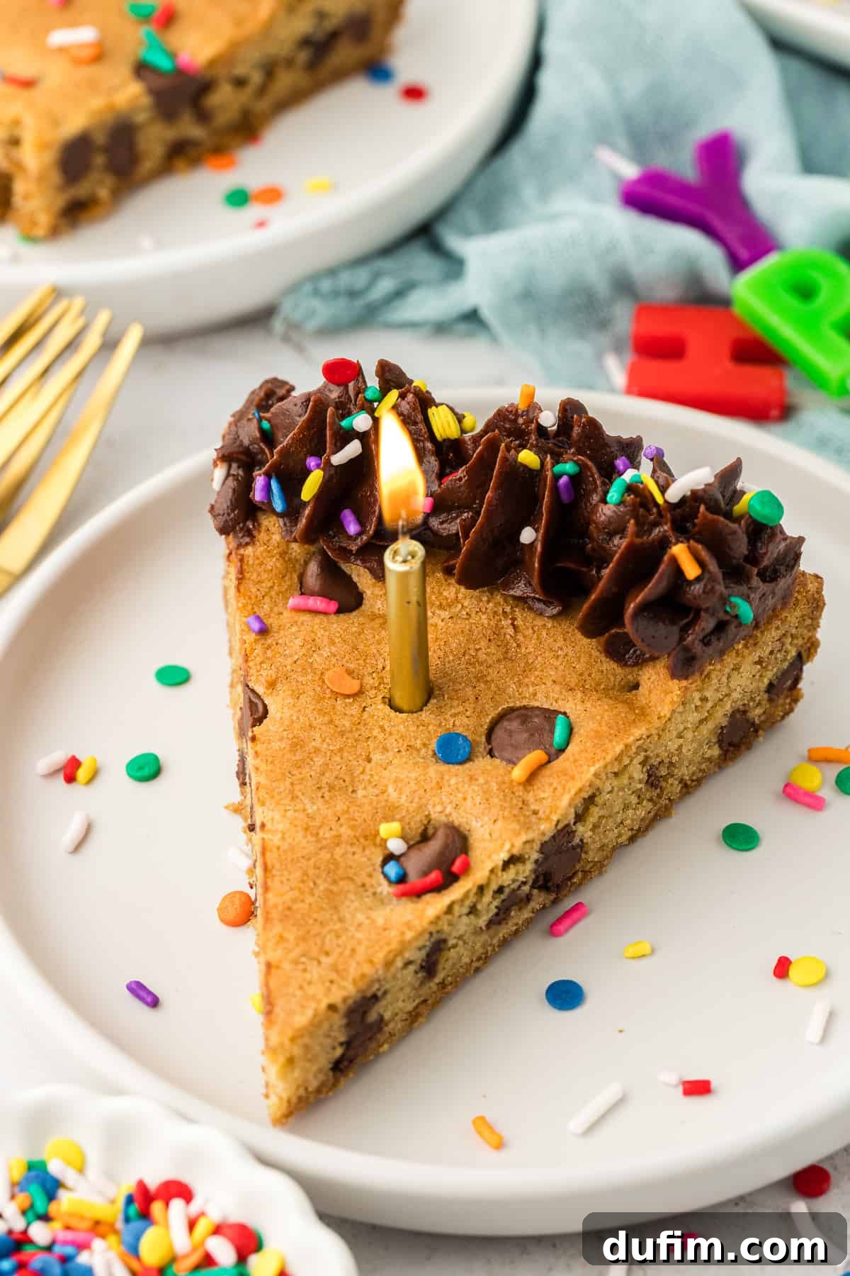 A festive table setting featuring a slice of chocolate chip cookie cake on a white plate, adorned with a single lit gold birthday candle, surrounded by additional gold forks, 'Happy Birthday' letter candles, and more plates holding delicious cake slices.