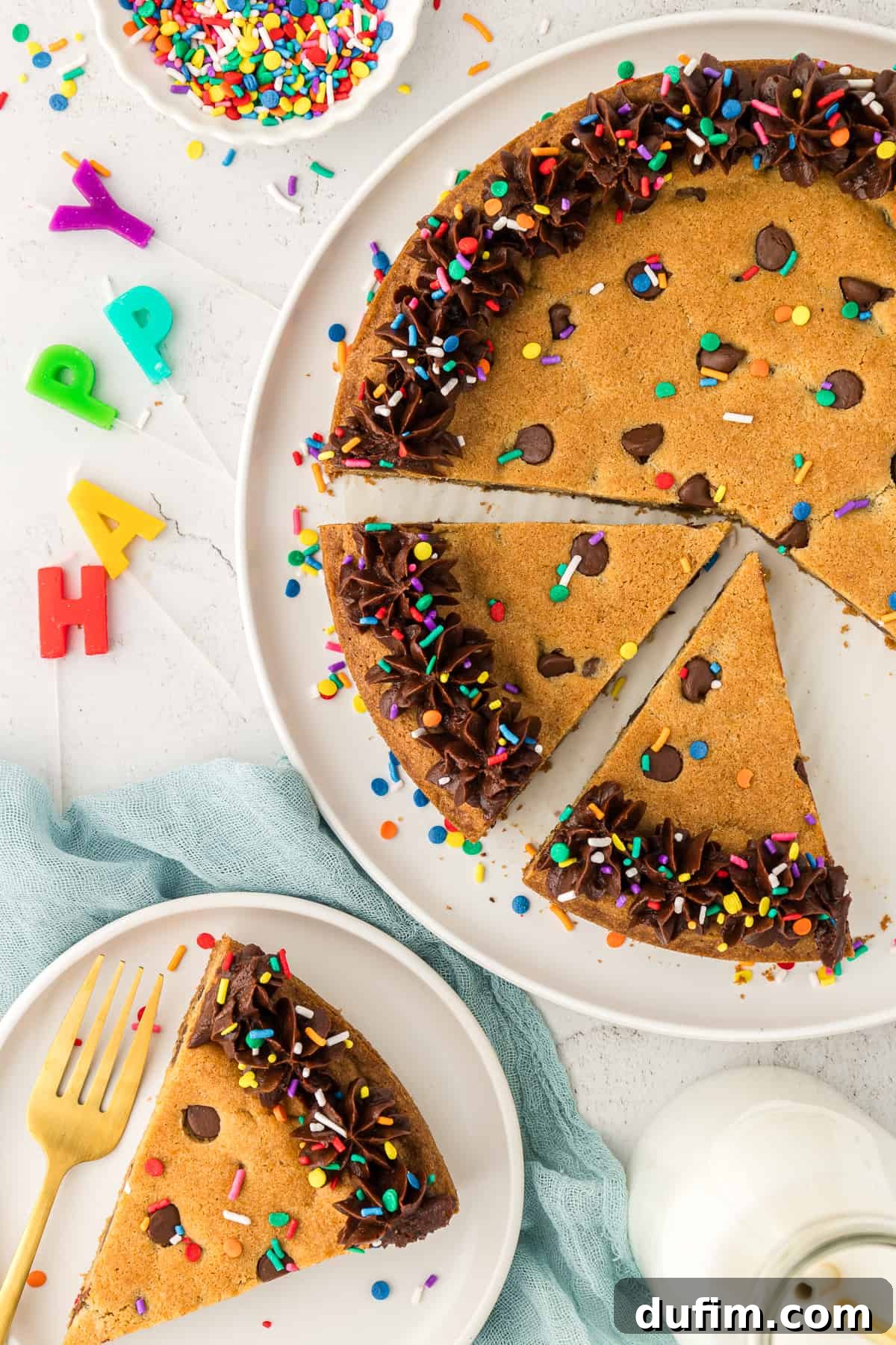 A festive display featuring a large chocolate chip cookie cake on a white plate with two slices already removed. Another slice rests on a smaller white plate with a fork and 'Happy Birthday' candles, accompanied by a bowl of colorful sprinkles.