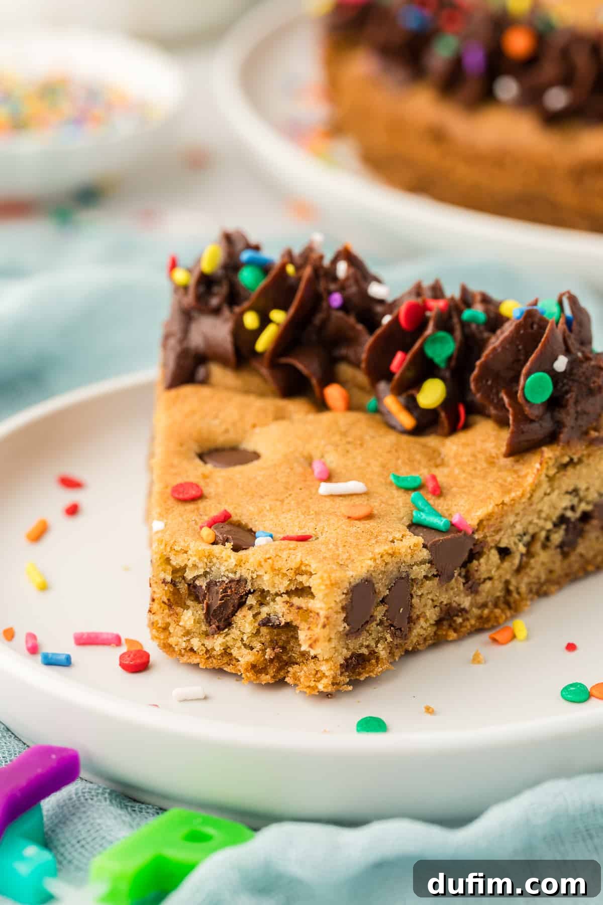 A mouth-watering slice of chocolate chip cookie cake on a white plate, beautifully decorated with chocolate frosting and festive colored sprinkles, with one tempting bite missing.