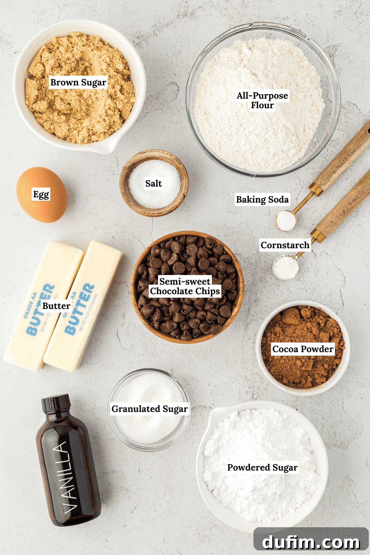 An inviting overhead shot of all the essential ingredients laid out for homemade cookie cake and chocolate frosting, including all-purpose flour, cornstarch, baking soda, salt, semi-sweet chocolate chips, softened unsalted butter, both brown and granulated sugars, a fresh egg, vanilla extract, unsweetened cocoa powder, and powdered sugar.