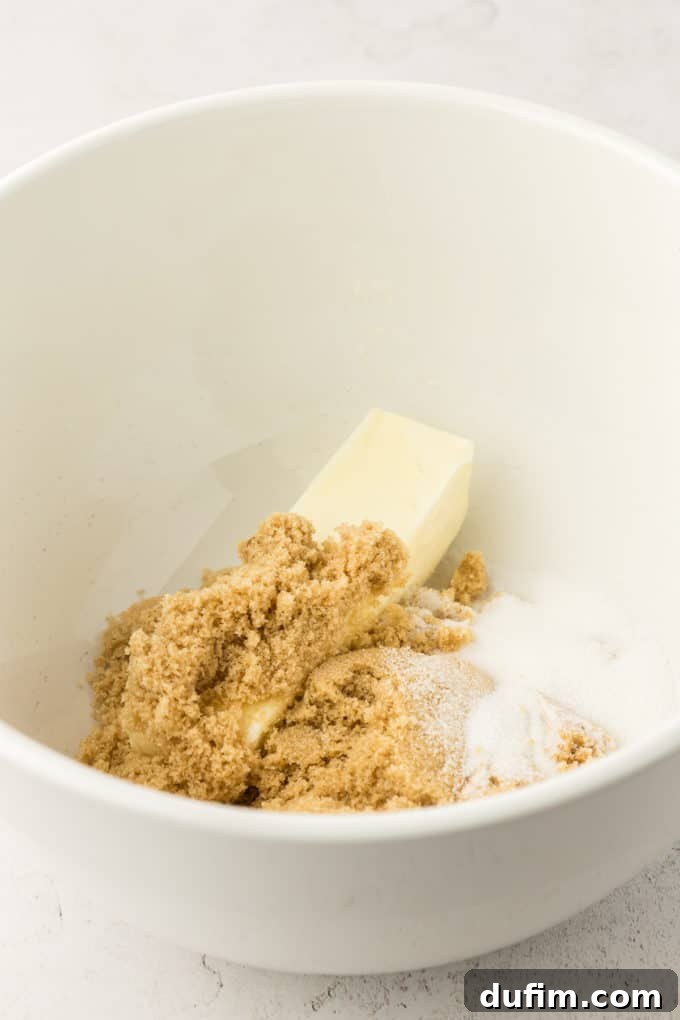 A stick of softened butter, rich brown sugar, and sparkling granulated sugar ready in a white mixing bowl, awaiting the creaming process.
