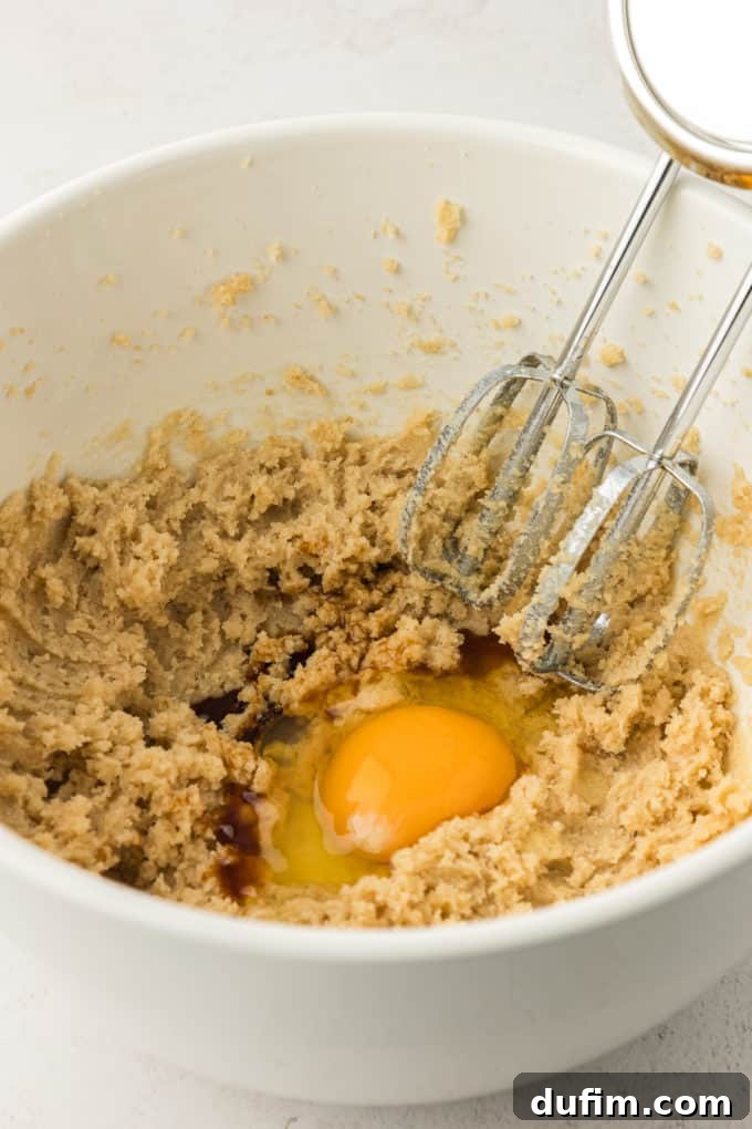The wet ingredients for the cookie cake, including butter and sugars, in a white bowl, with an electric mixer resting, showing the fluffy, pale mixture after creaming.