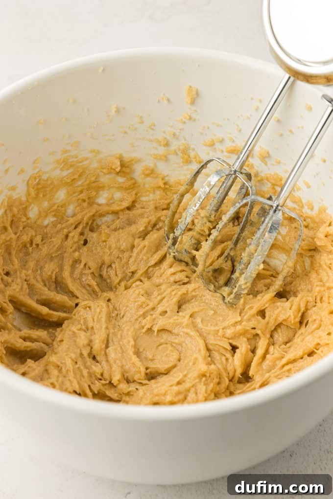 Another angle of the creamy wet ingredients for the cookie cake in a white bowl, highlighting the light and airy texture achieved by the electric mixer.