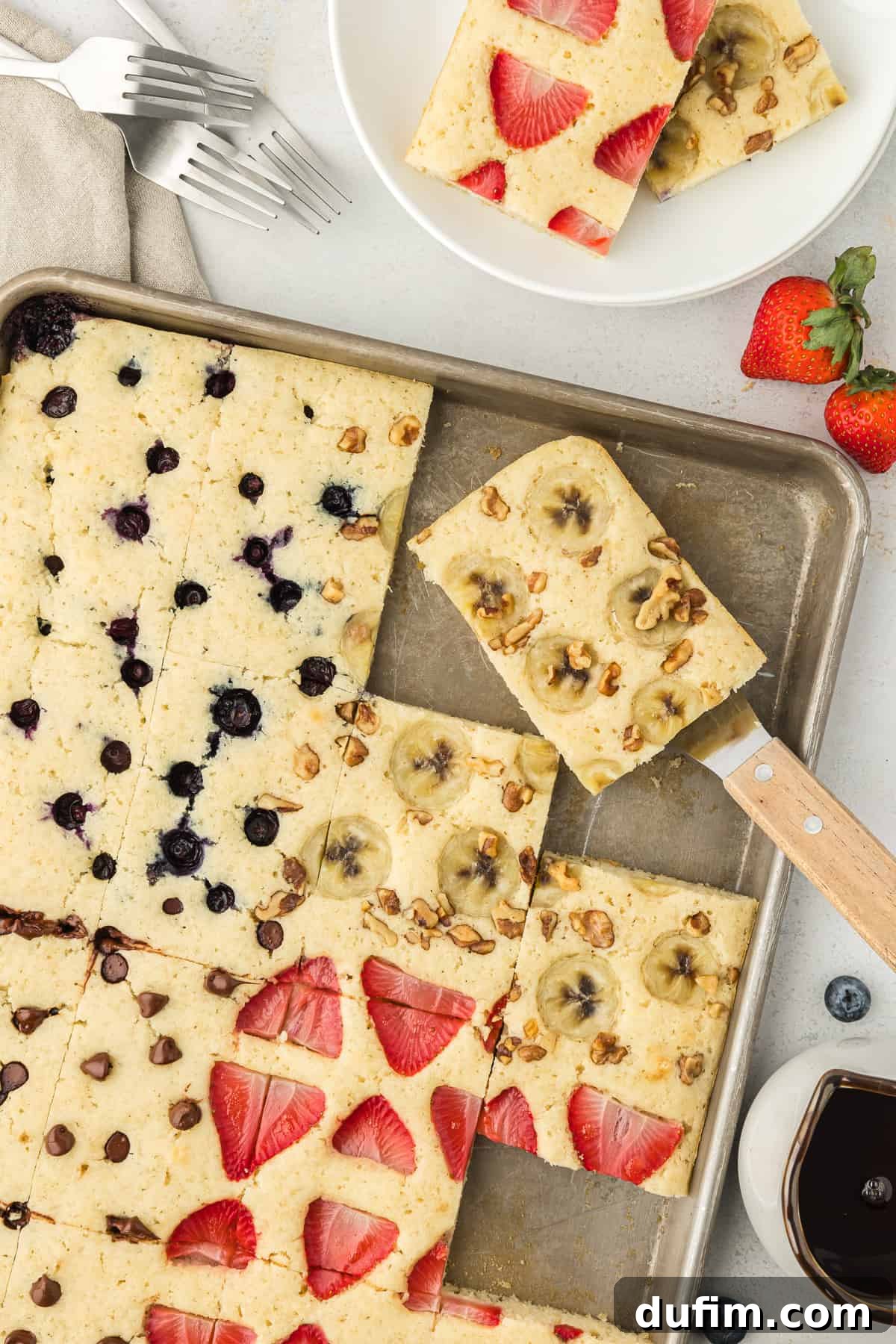A sheet pan full of pancakes, some with blueberries in them, some with bananas and nuts, some with chocolate chips and some with strawberries, with one pancake being lifted by a spatula, syrup, fresh blueberries and strawberries, forks, napkins, and a plate with two slices of pancake around the sheet pan