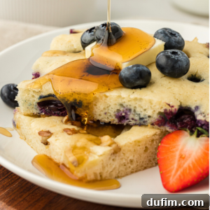 two slices of sheet pan pancakes on a white plate topped with fresh blueberries, a slice of butter and being drizzled with syrup, with a fresh strawberry slice on the plate beside the pancakes