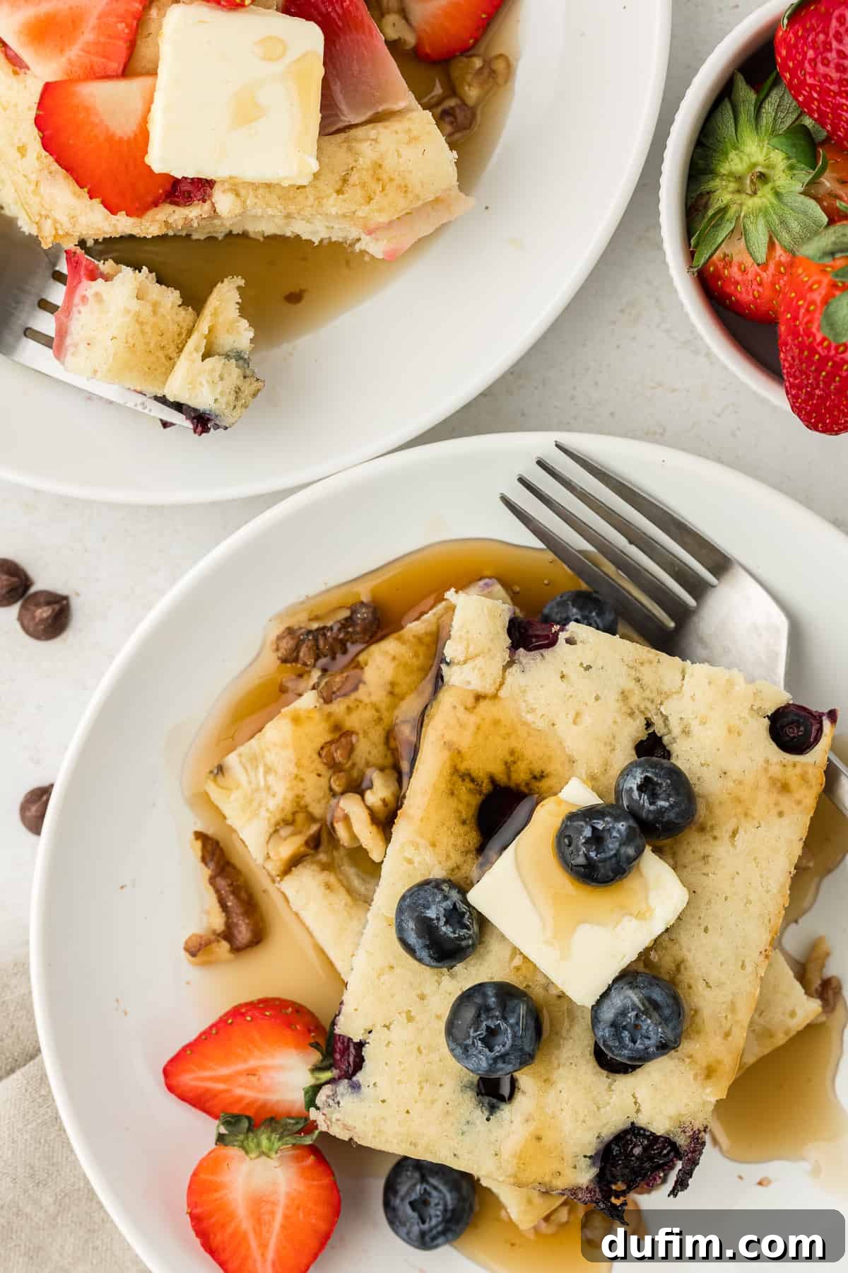 two white plates with slices of sheet pan pancakes on them, topped with slices of butter, maple syrup and fresh blueberries and strawberries, with forks on both plates and a bowl of fresh strawberries beside them