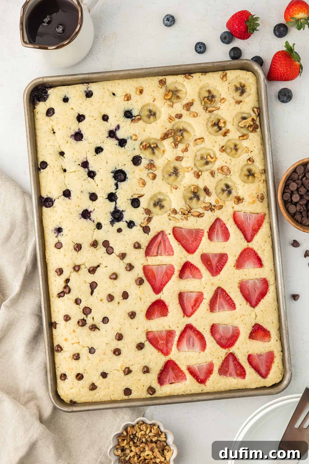 A sheet pan full of pancakes, blueberry pancakes in the upper left corner, banana walnut pancakes in the upper right corner, chocolate chip pancakes in the lower left corner and strawberry pancakes in the lower right corner, with syrup, fresh berries, a cup of chocolate chips, a cup of chopped nuts and a light tan towel surrounding the sheet pan