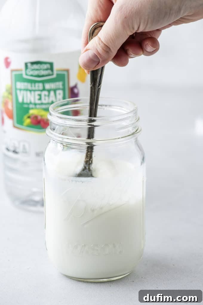 a clear glass jar of buttermilk being stirred with a spoon, with a bottle of white vinegar in the background