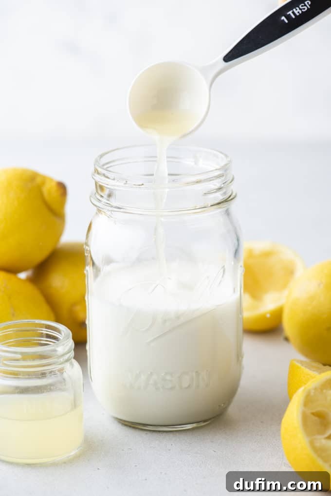 a mason jar of buttermilk with a tablespoon of lemon juice being poured in, a small clear glass jar of lemon juice beside it, surrounded by fresh whole and sliced lemons