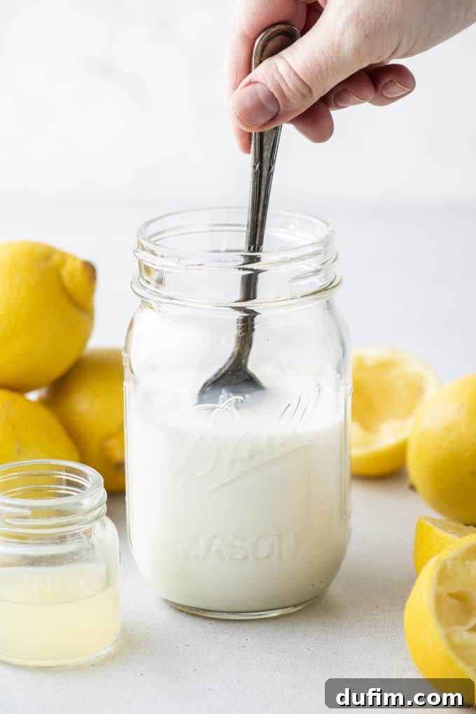a mason jar with buttermilk in it being stirred, a small clear glass jar of lemon juice beside it and whole and sliced lemons surrounding it