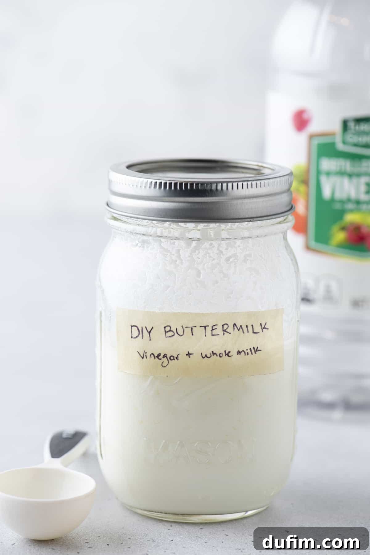 A mason jar full of buttermilk with a lid on, labeled with tape that says "DIY Buttermilk: vinegar + whole milk", with a measuring spoon beside it an a bottle of vinegar in the background