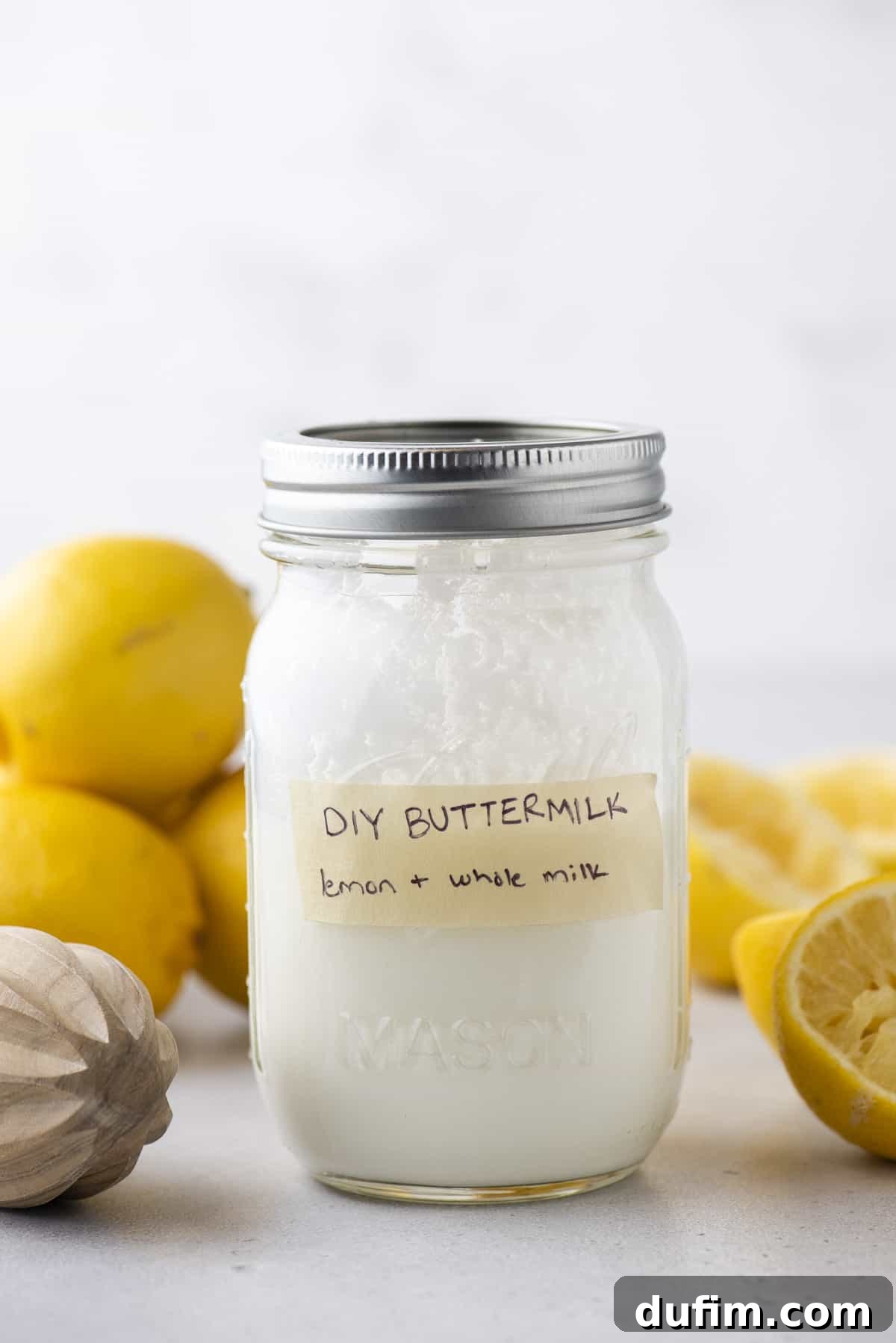 A mason jar full of buttermilk with a strip of tape on it labeled "DIY Buttermilk: lemon + whole milk", with fresh lemons all around