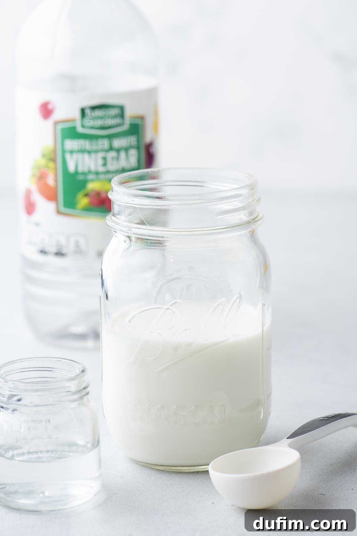 A clear glass jar of buttermilk with a white tablespoon and a small clear glass jar of vinegar and a bottle of vinegar in the background