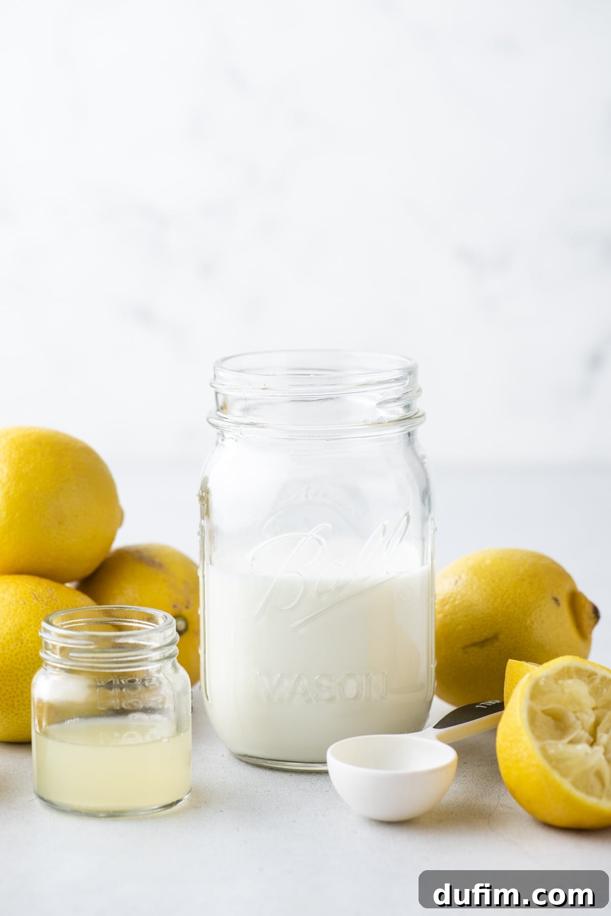 A mason jar full of buttermilk surrounded by fresh whole and sliced lemons, a white tablespoon measurer and a small clear glass jar of lemon juice