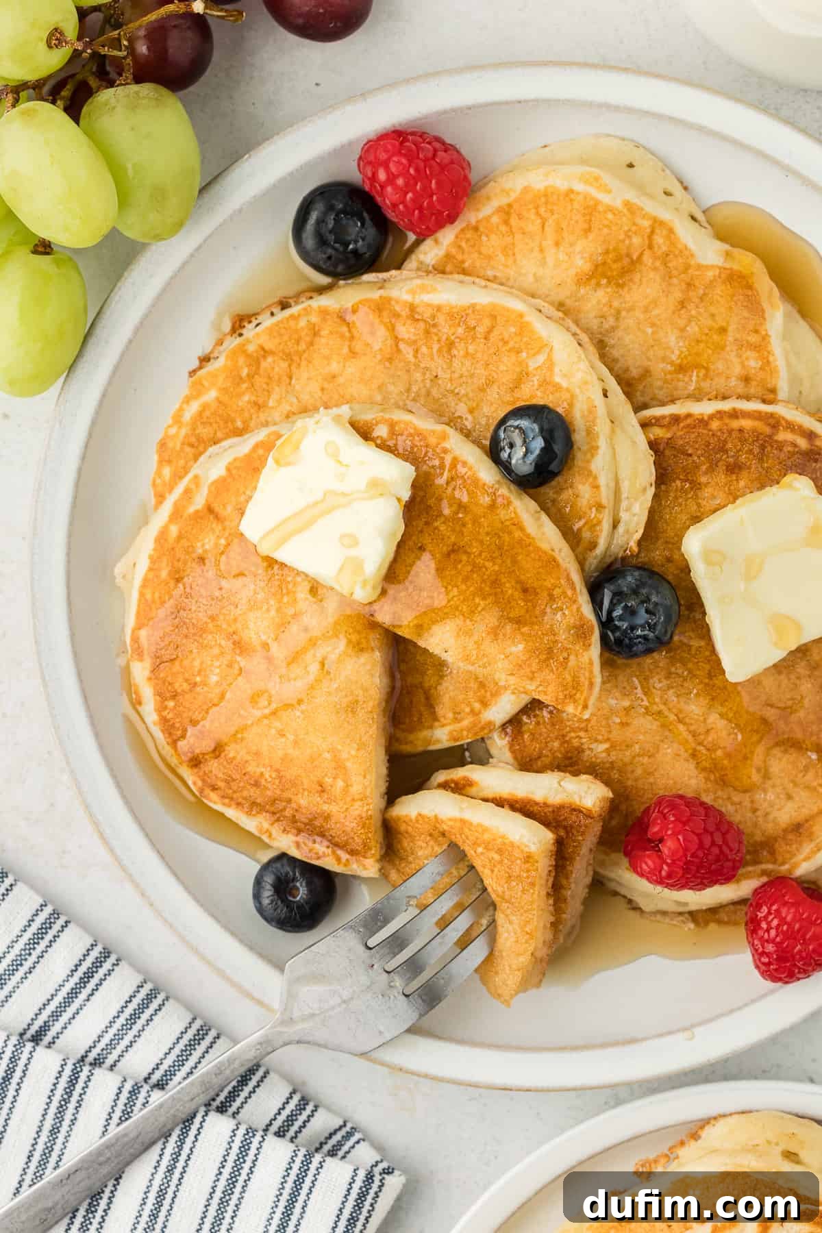 Simple Secrets to Fluffy Buttermilk Pancakes 2 A white plate showcasing a stack of four golden-brown buttermilk pancakes, generously topped with fresh blueberries, raspberries, pats of melting butter, and a glistening drizzle of maple syrup. A fork holds a triangle-shaped bite from two pancakes, poised above the plate, hinting at their soft texture. The scene is completed with a blue and white striped towel, fresh grapes, and another plate of pancakes in the background.