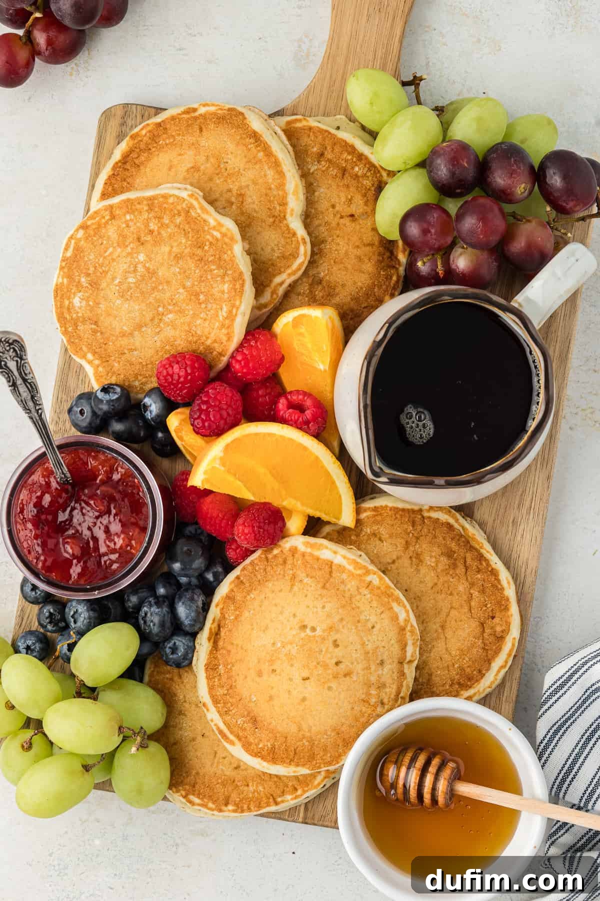 Simple Secrets to Fluffy Buttermilk Pancakes 11 A rustic wooden cutting board overflowing with a bounty of fluffy buttermilk pancakes, accompanied by a selection of delectable toppings including a bowl of rich maple syrup, a small jar of vibrant fruit jam with a spoon, a pot of golden honey, and an array of fresh fruits like juicy orange slices, plump raspberries, sweet blueberries, and vibrant green and red grapes, creating a visually stunning breakfast spread.