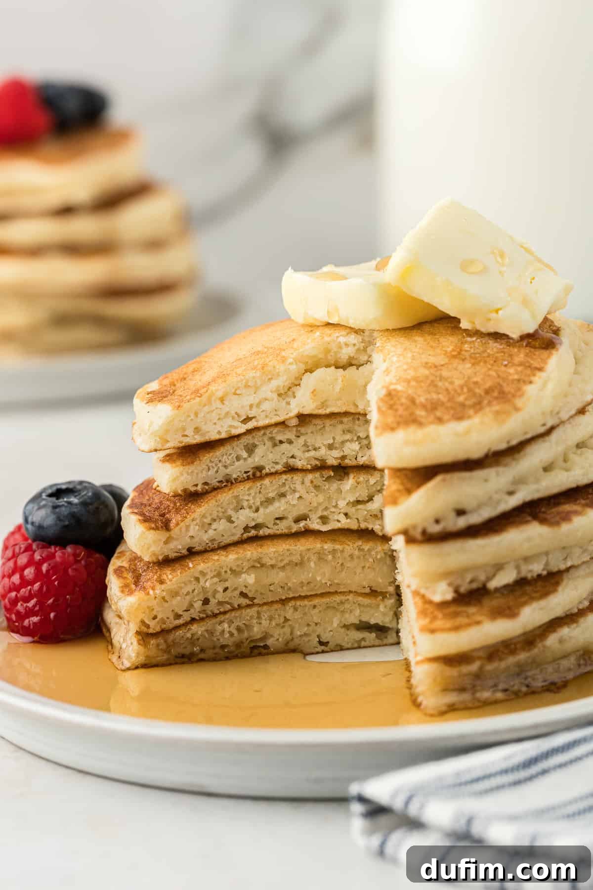 Simple Secrets to Fluffy Buttermilk Pancakes 12 A white plate holding a towering stack of five perfectly golden buttermilk pancakes, with a large, inviting slice removed from the entire stack. The plate is adorned with fresh blueberries and raspberries, two pats of melting butter atop the pancakes, and abundant drizzles of glistening maple syrup creating pools around the stack.